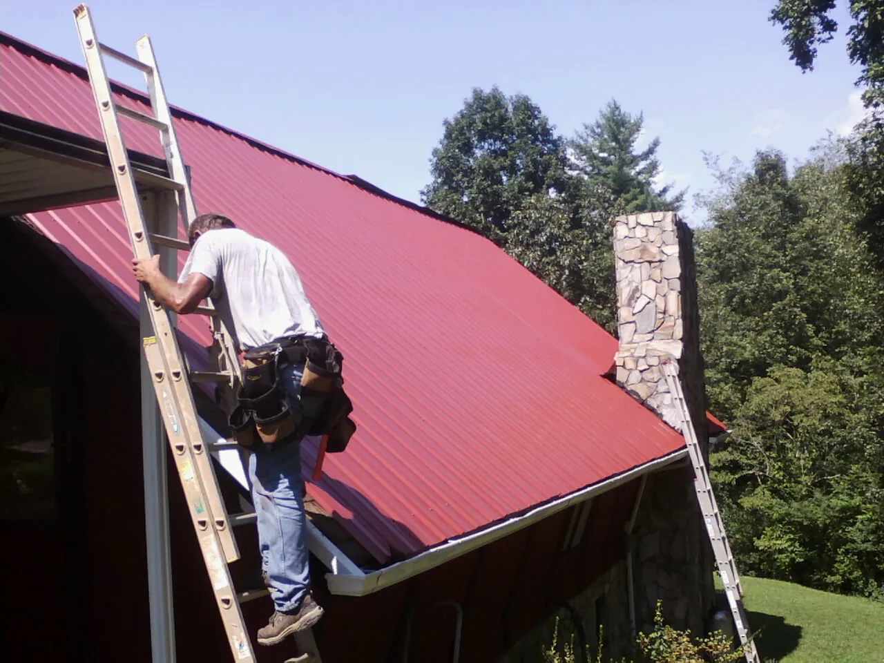 Roofer on a ladder beside a red metal roof and stone chimney — Soffit & Fascia Repair by Roofing Brothers in Amesville
